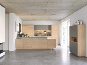 Fußboden in Betonoptik mit Einbauküche aus Naturholz in Eiche. Blick über die Kücheninsel zur Küchenzeile und einem Küchenregal. 